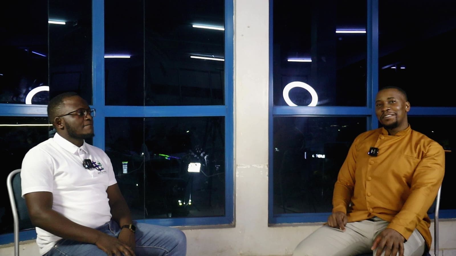 Two people seated on the podcast set, ring lights reflected in the glass and city lights beyond