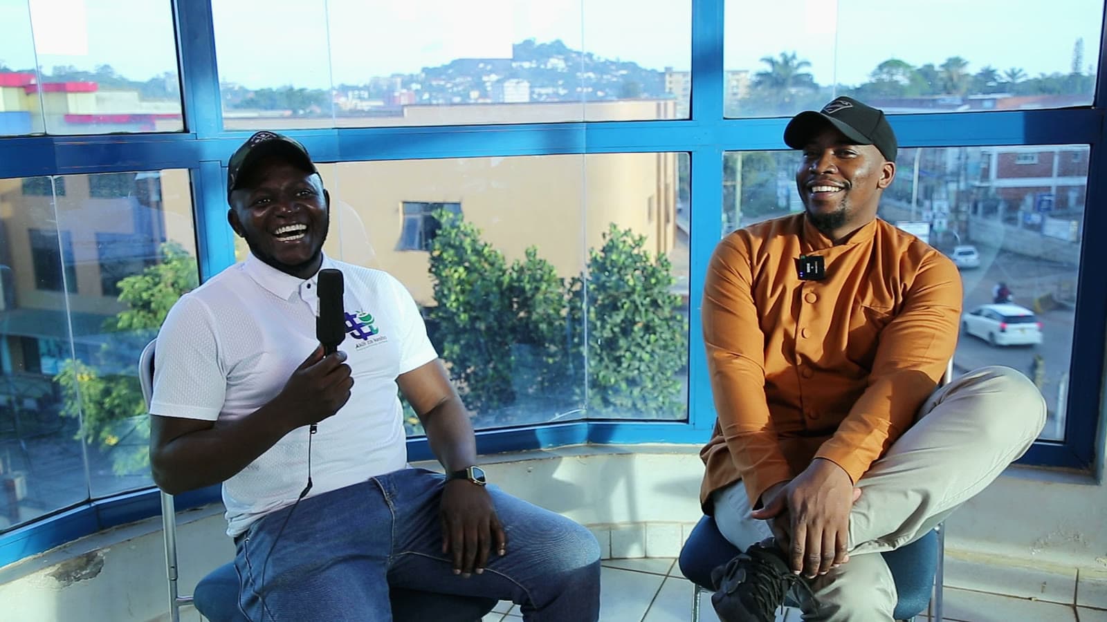 Daytime on set: two people laughing, the city skyline behind the glass
